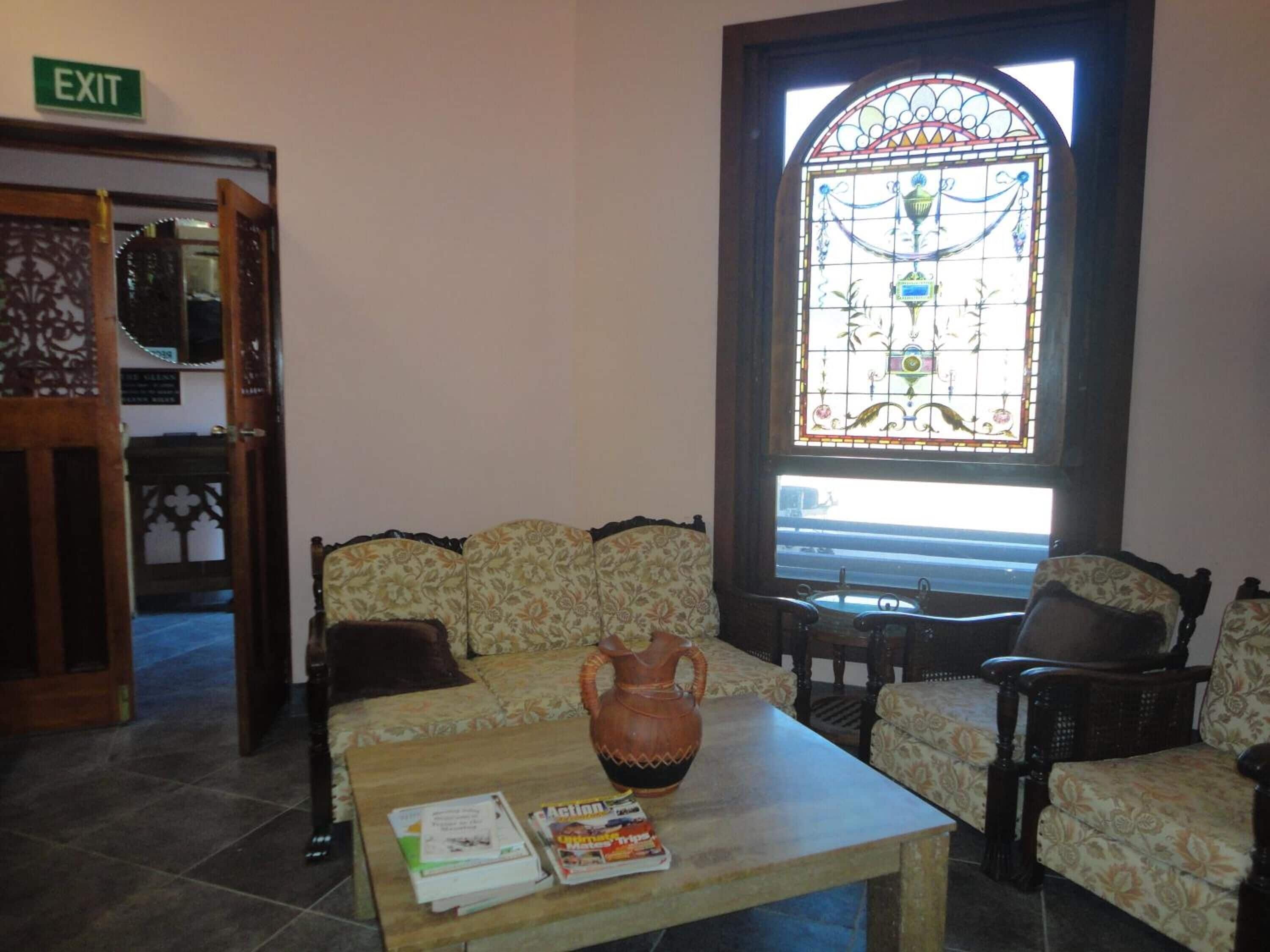 A lobby with a sofa, chairs, a coffee table with books, a large stained glass window, and an exit sign.