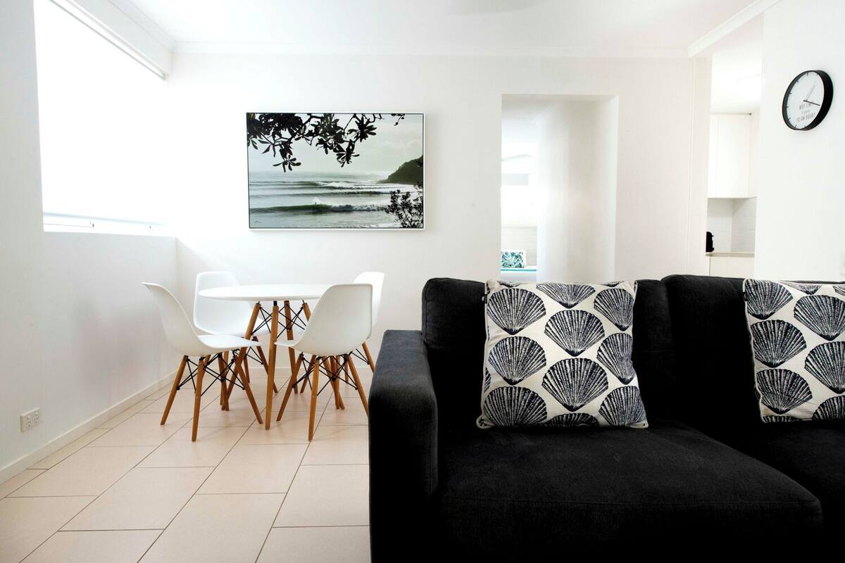 Een moderne woonkamer met een zwarte bank, witte stoelen en een eettafel. Op de muur hangt een schilderij van een strandgezicht.