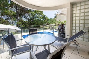 A sunlit outdoor patio with a round table and chairs, a pool, and a view of trees and a building in the background. - Sandcastles Noosa (Noosa Heads)