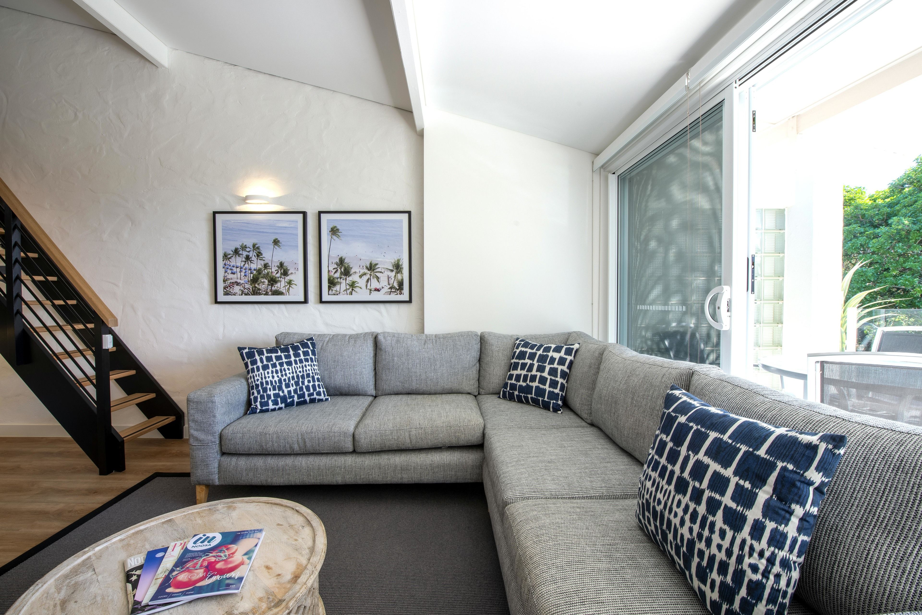 A modern living room with a grey sectional sofa, two patterned throw pillows, a coffee table with magazines, and a staircase.