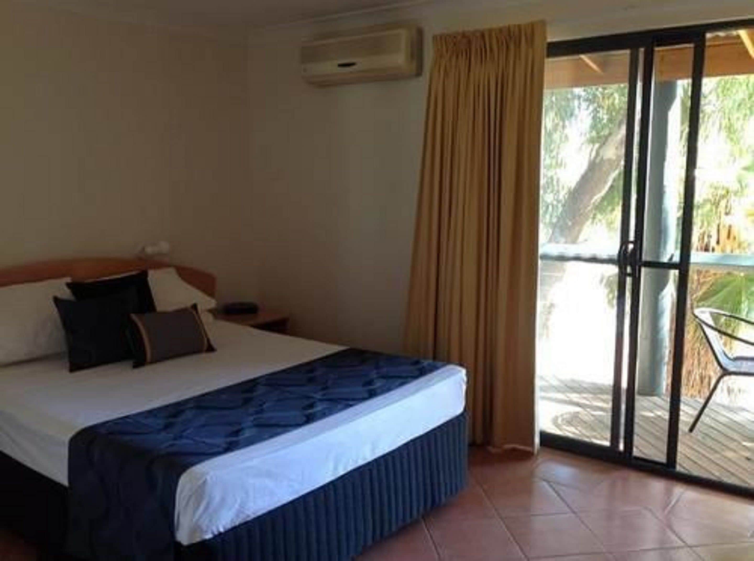 A hotel room with a bed, a nightstand, a window with curtains, and a view of the outdoors.
