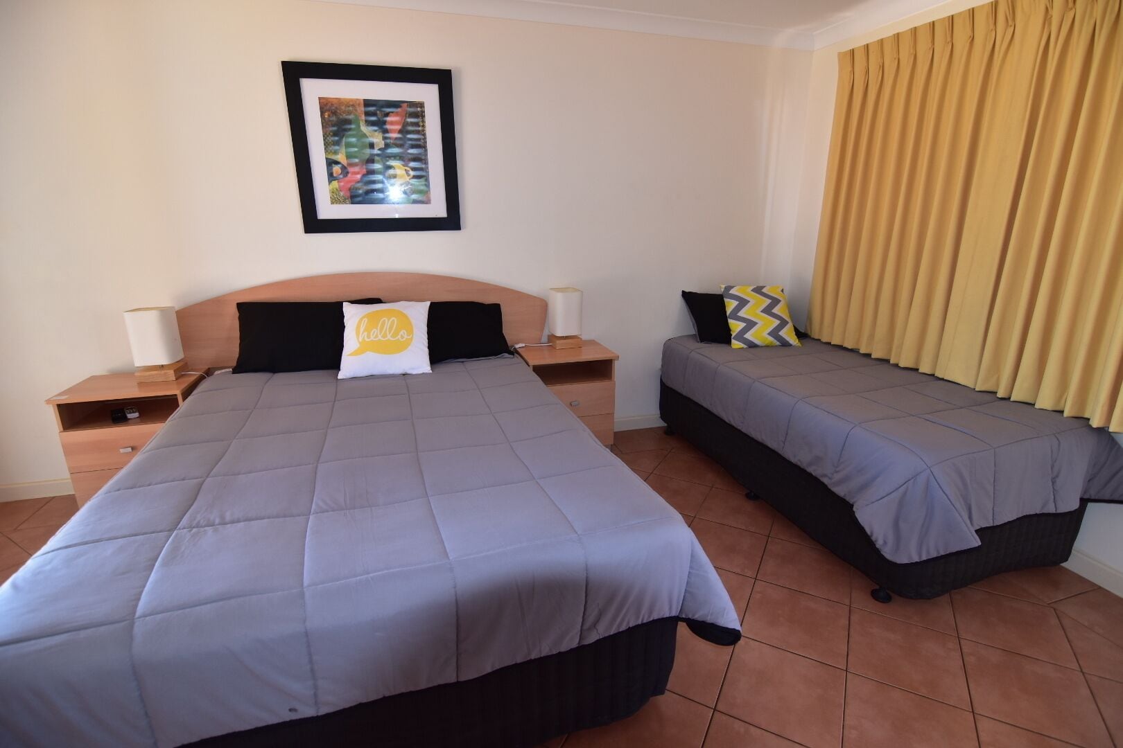 A hotel room with two beds, a nightstand with a lamp, a framed picture on the wall, and a window with curtains.