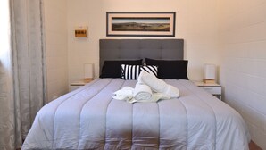 Una cama bien hecha con colcha gris, almohadas a rayas negras y blancas y toallas enrolladas sobre la cama. Encima de la cama hay un cuadro enmarcado, una mesita con una lámpara y un reloj de pared.