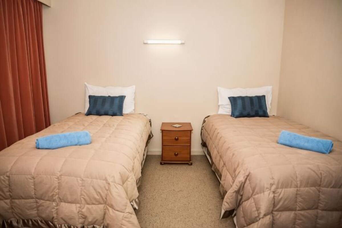 A twin room with two beds, each with a blue pillow and a beige blanket. There is a wooden nightstand between the beds with a small drawer.