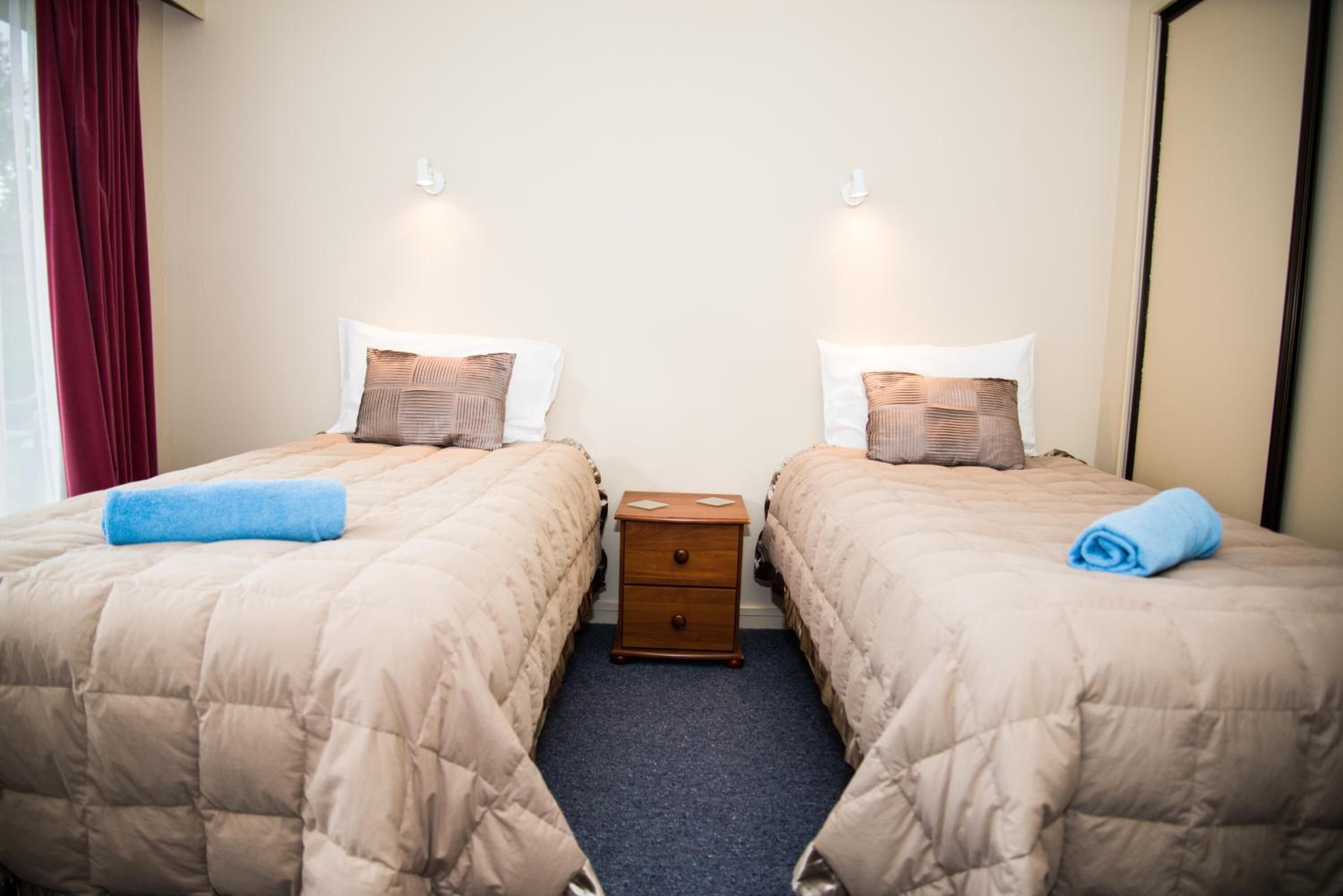 Een hotelkamer met twee bedden, elk voorzien van een beige gebogen sprei, kussens en een blauwe handdoek. Daartussen staat een houten nachtkastje met een klein lade.