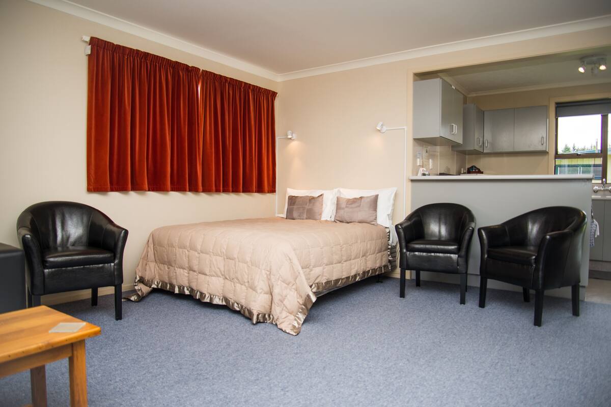 A bedroom with a bed, two chairs, a table, and a kitchen area visible through an open door.