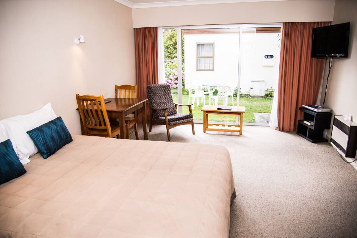 A hotel room with a large bed, a wooden table with chairs, a television, and a sliding glass door leading to an outdoor area with chairs and a table.