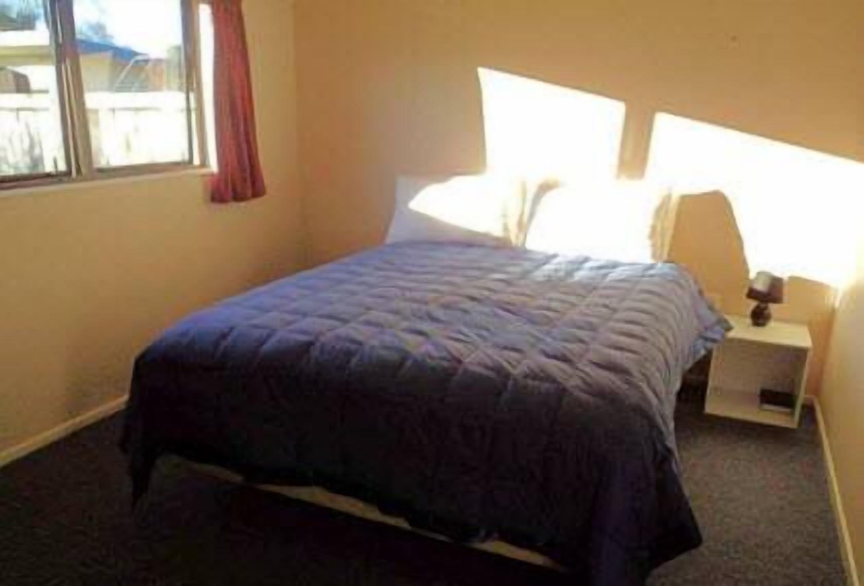 Un dormitorio con cama, mesita de noche, lámpara y ventana con cortinas.