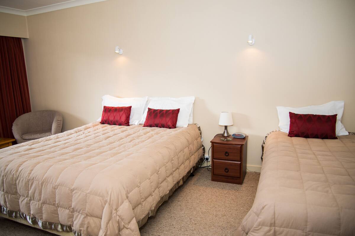 A hotel room with two beds, each with a beige quilted bedspread and red pillows. There is a wooden nightstand with a lamp between the beds.