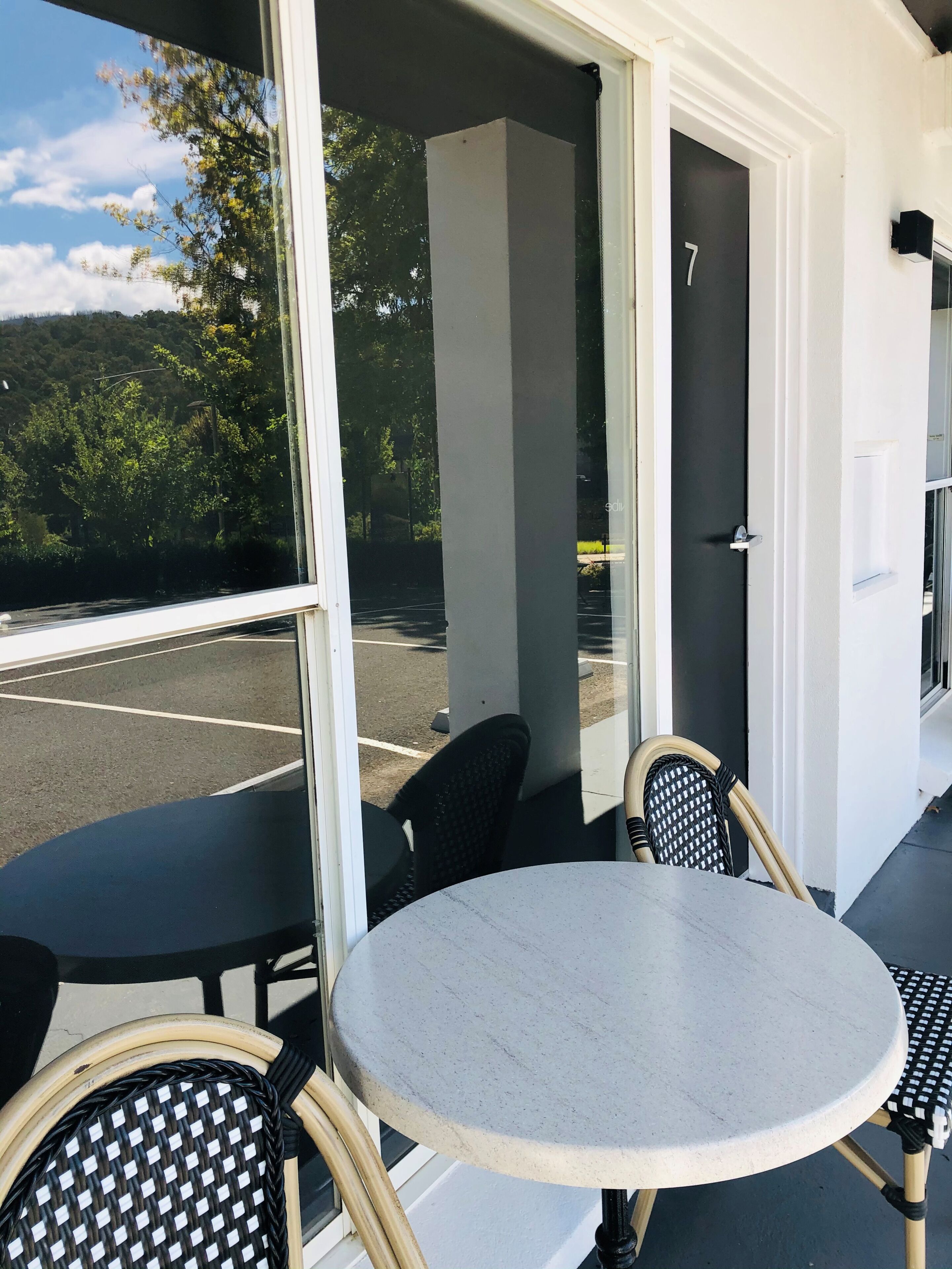 Una stanza con un tavolo rotondo e due sedie, una porta con il numero 7, e vista su un parcheggio e alberi.