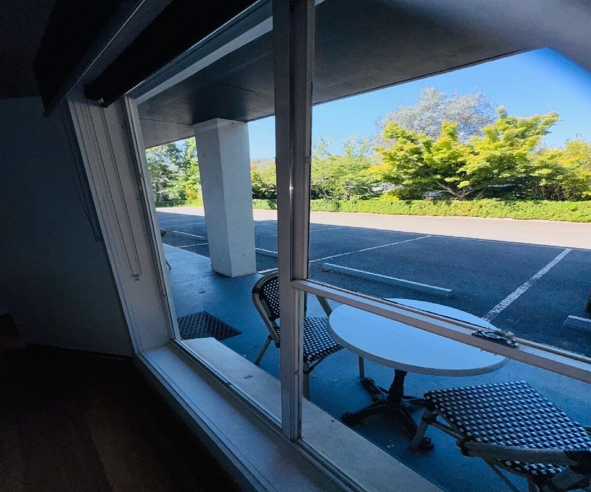 A view from a window with a table and chairs outside, a parking lot, and greenery in the background.