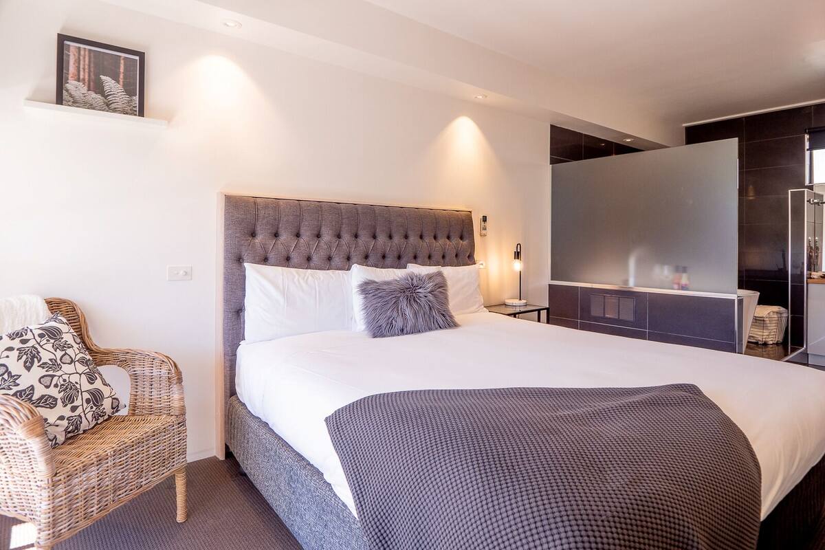 Una habitación de hotel con una cama grande, una silla de mimbre, una mesita de noche con lámpara y un espejo grande.