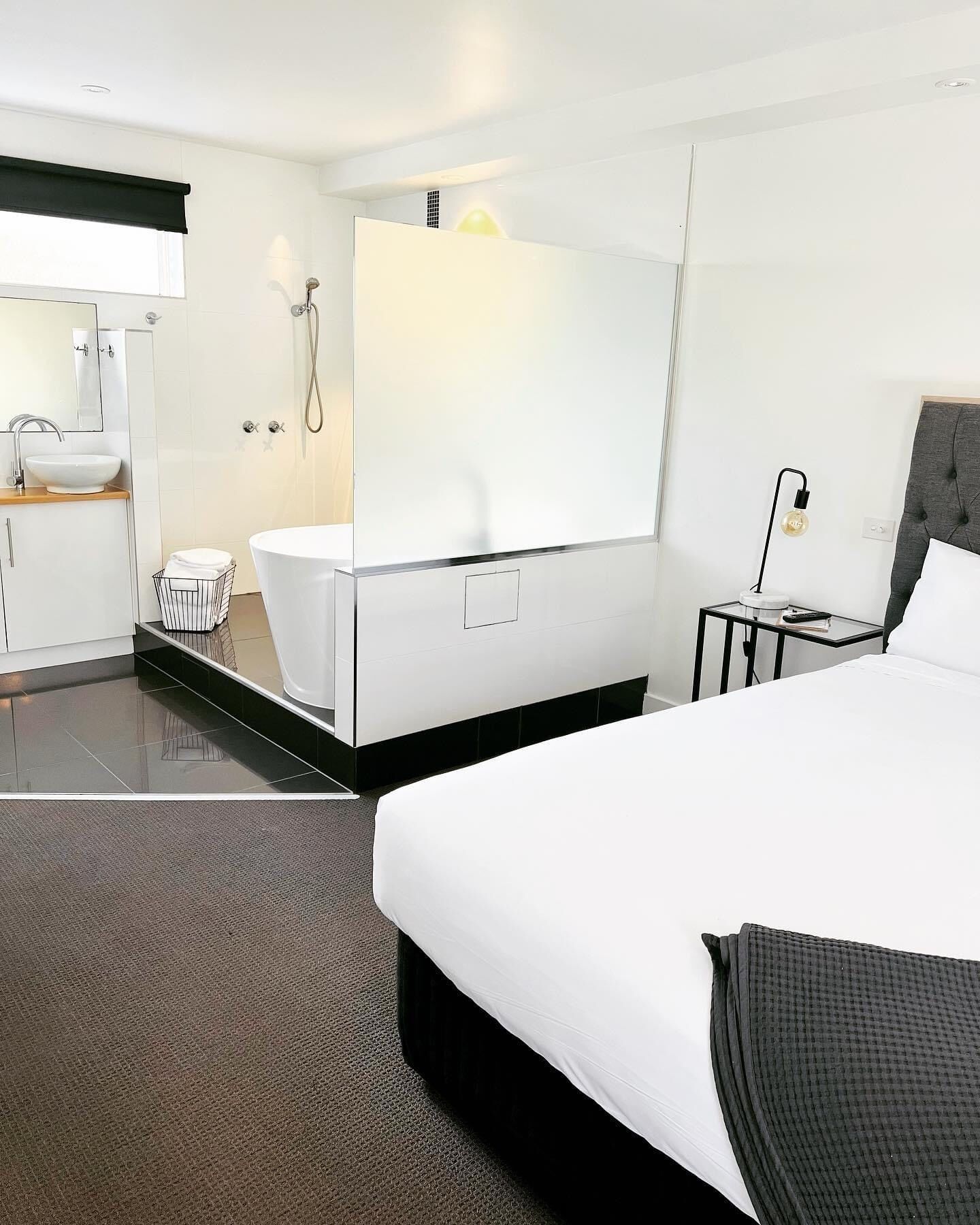 A modern hotel room with a large bed, a separate bathroom with a bathtub, a small bedside table with a lamp, and a large mirror above the mirror.