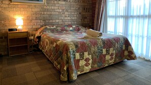 A bedroom with a brick wall, a painting, a bedside lamp, a quilted bedspread, and a window with curtains.