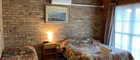Una habitación con dos camas, una pared de ladrillo, un cuadro, una lámpara y una ventana con cortinas.