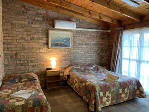 A room with two beds, a brick wall, a painting, a lamp, and a window with curtains.