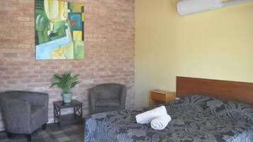 Quarto de hotel com uma cama, duas poltronas, uma mesinha com uma planta e um quadro numa parede de tijolos.