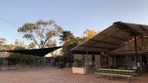 An outdoor seating area with picnic tables and a shaded area, surrounded by trees and a building with signage.