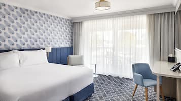 A hotel room with a large bed, a desk, a chair, a small table, and a window with curtains.