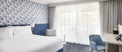 A hotel room with a large bed, a desk, a chair, a small table, and a window with curtains.