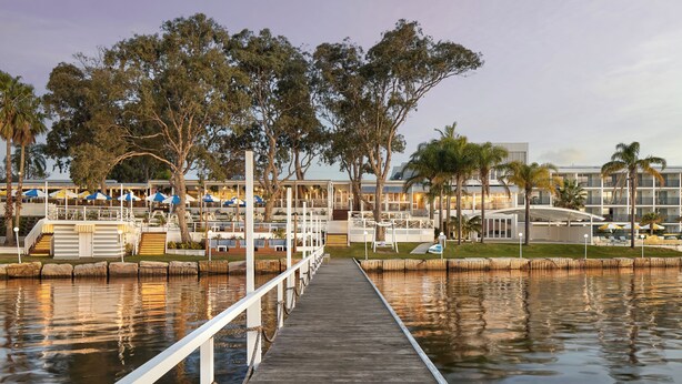 A waterfront hotel with a wooden dock leading to a pool area, surrounded by trees and outdoor seating.