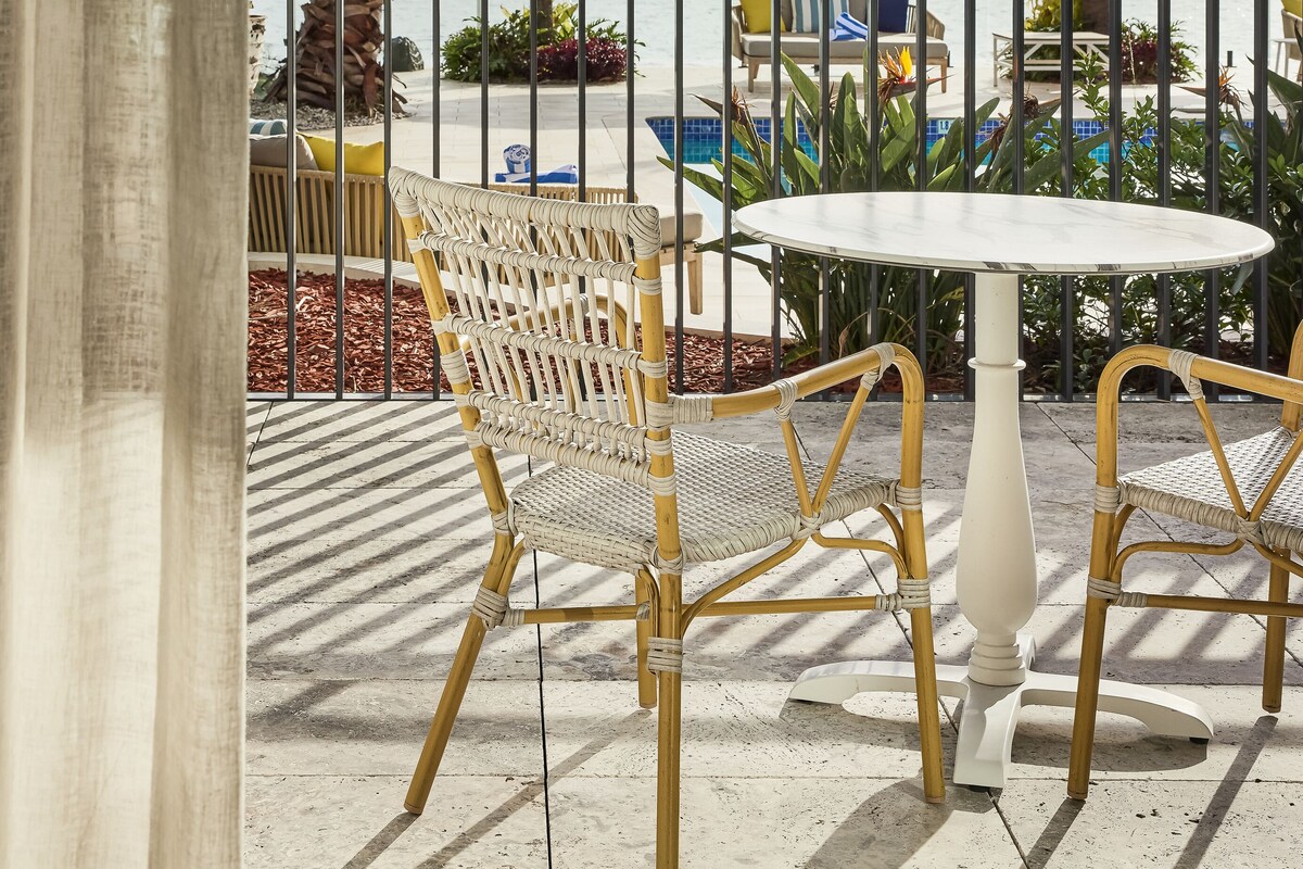 A balcony with wicker chairs and a round table, overlooking a pool and palm trees.