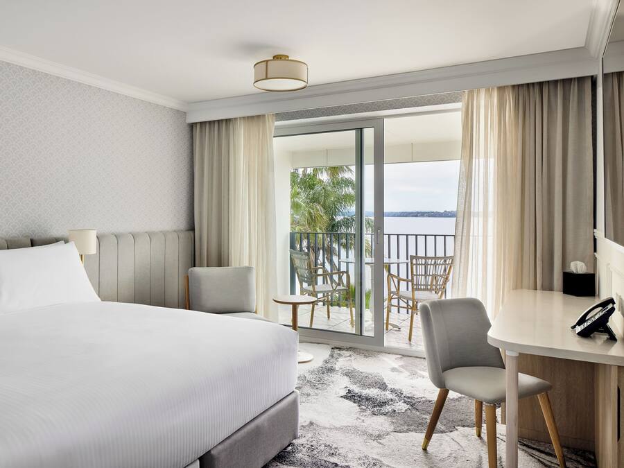 A hotel room with a large bed, a desk with a chair, a balcony with a view of the water, and a patterned wall.