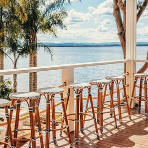 A wooden deck with six wicker bar stools facing a body of water with a clear sky. - The Beachcomber Hotel & Resort (Toukley)