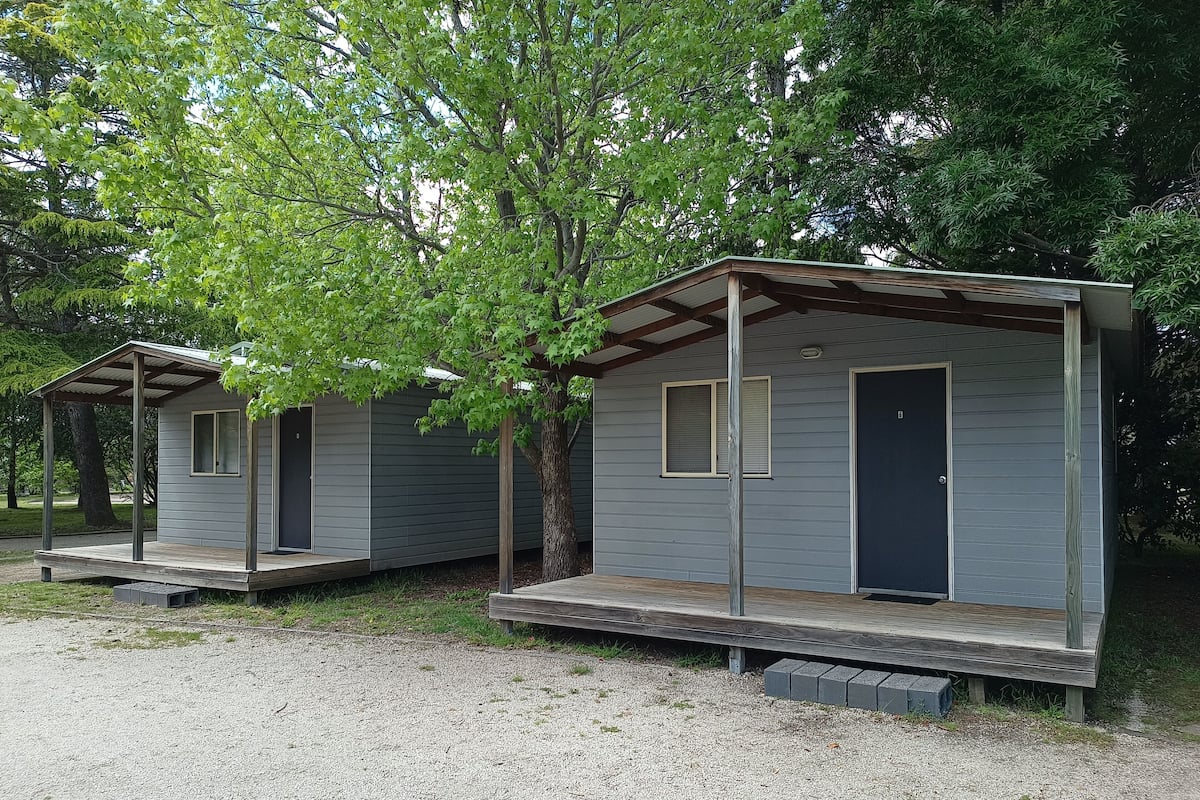 Two small, single-story cabins with a porch, surrounded by trees.