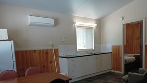 A compact kitchen with a dining table, chairs, a refrigerator, a window with blinds, and a wooden wall panel. - Lithgow Tourist and Van Park (Bowenfels)