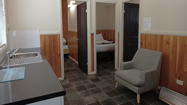 A compact room with a kitchenette, a grey armchair, and a bed with wooden paneling. - Lithgow Tourist and Van Park (Bowenfels)