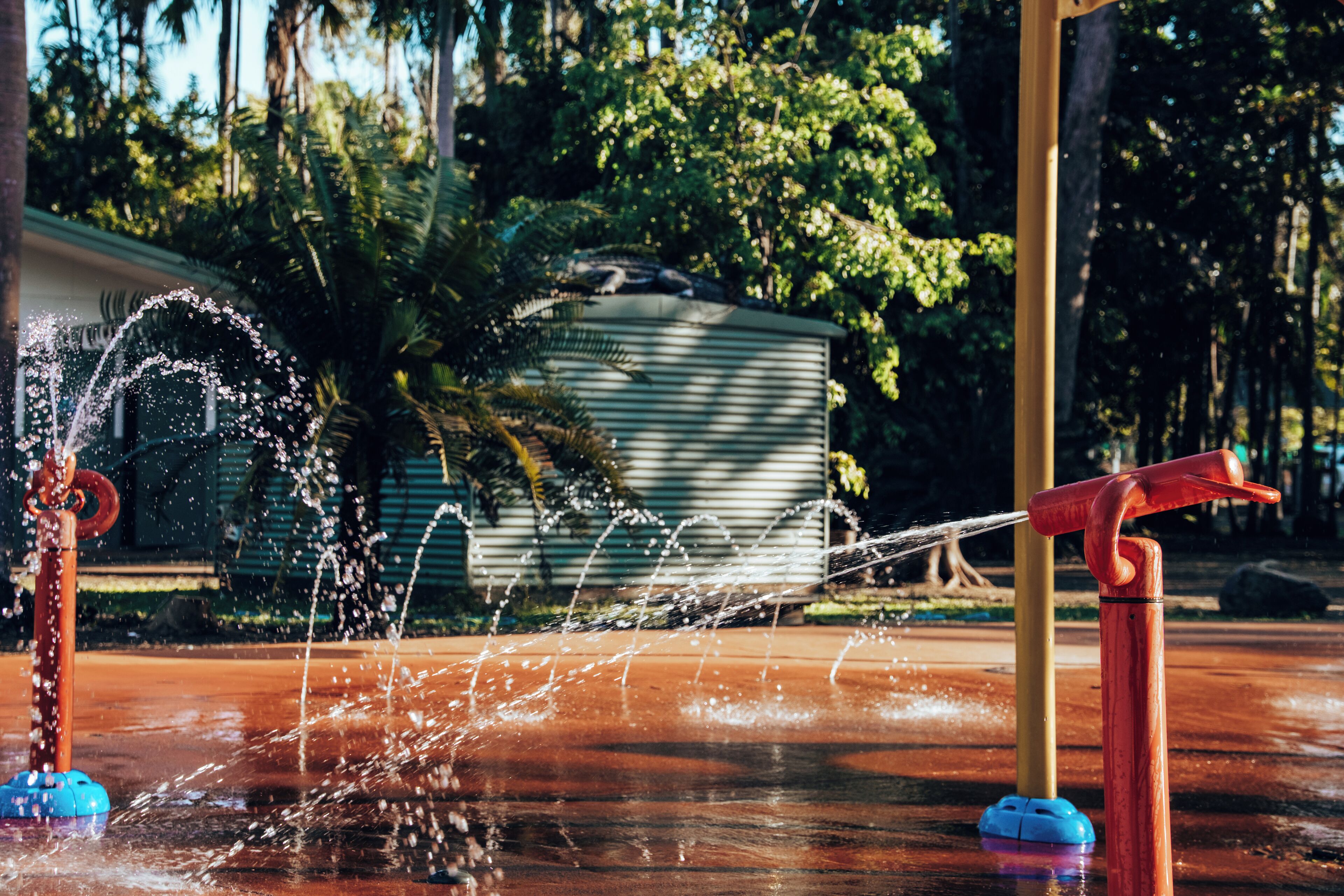 A water feature with a fountain and a water slide, surrounded by greenery and a building in the background.