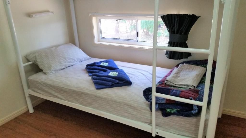 Una cama individual con ventana encima, un estante pequeño y una cortina.