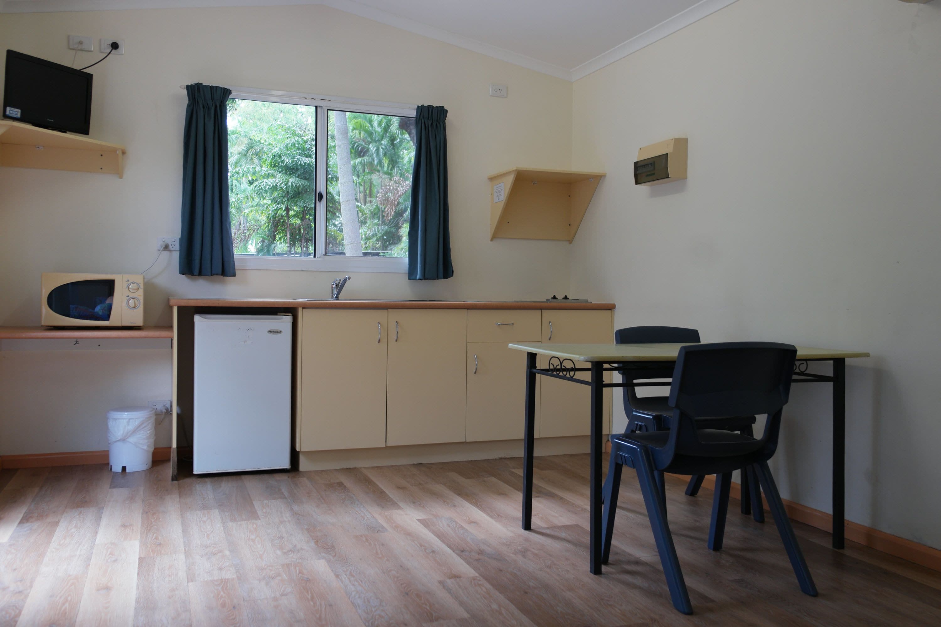 A compact kitchen with a microwave, refrigerator, and a small sink. There is a dining table with two chairs and a window with curtains.