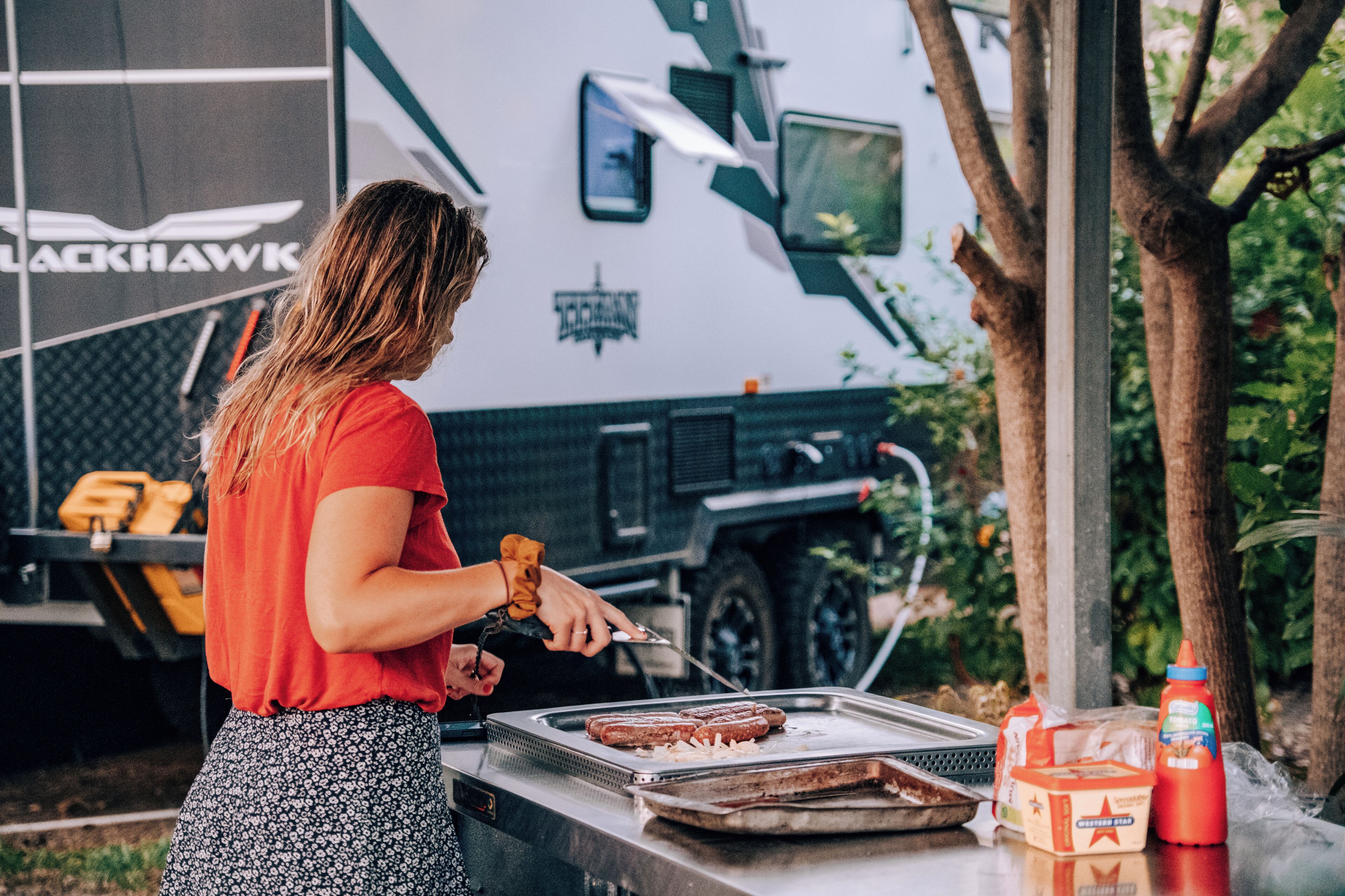 En person griller pølser udendørs, med en autocamper i baggrunden.