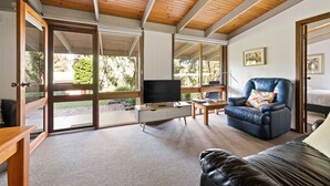 Uma sala de estar ampla com uma grande porta de vidro deslizante que dá para uma área externa, um sofá de couro, uma televisão sobre um móvel branco e teto de madeira.