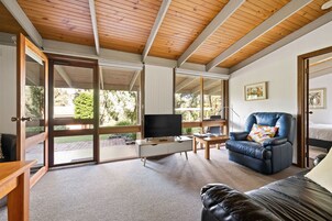 Un salón amplio con un gran ventanal corredera que da a un área exterior, un sofá de cuero, un televisor sobre un mueble blanco y un techo de madera.