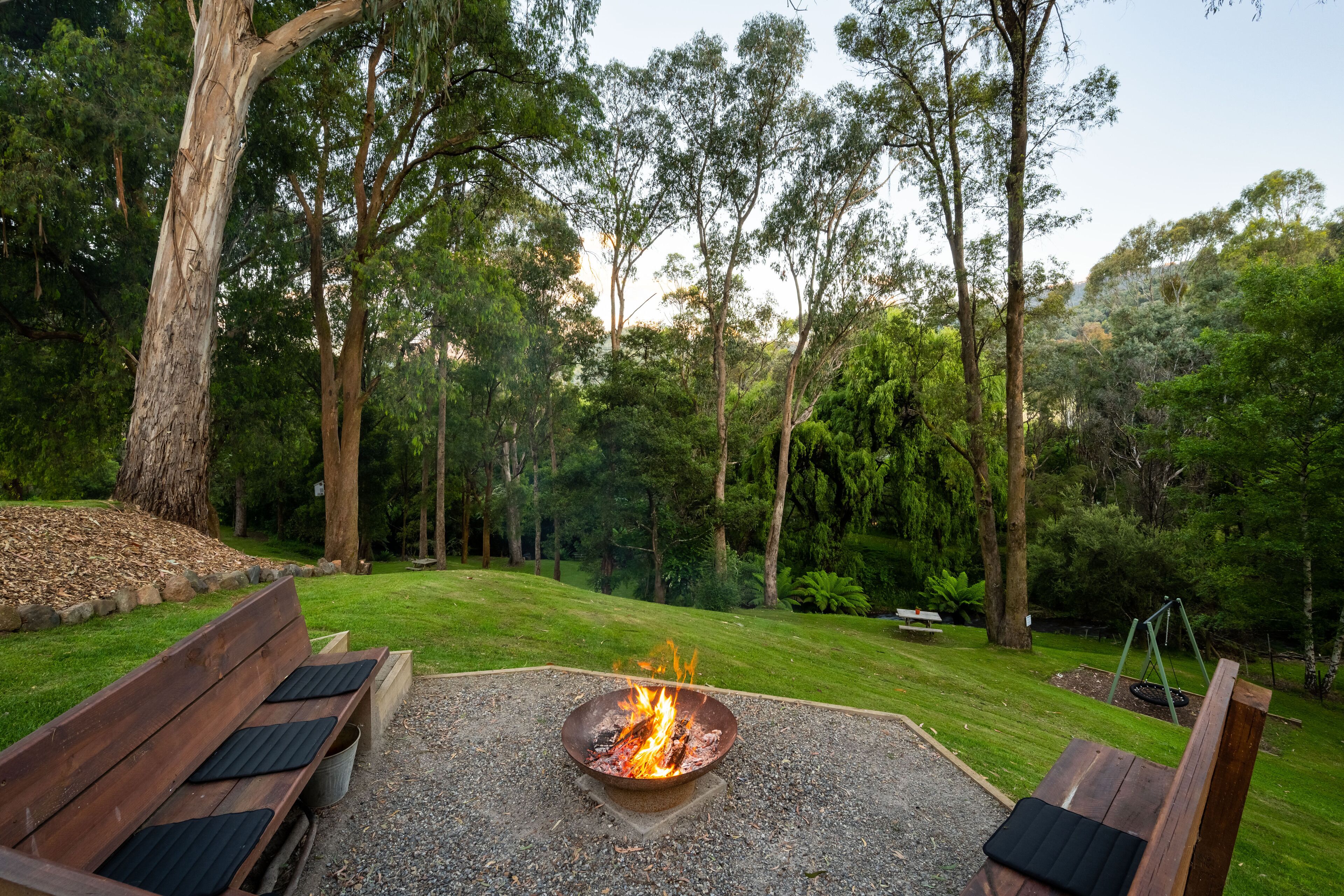 A fire pit surrounded by a wooden bench and a swing set in a lush, green outdoor area.