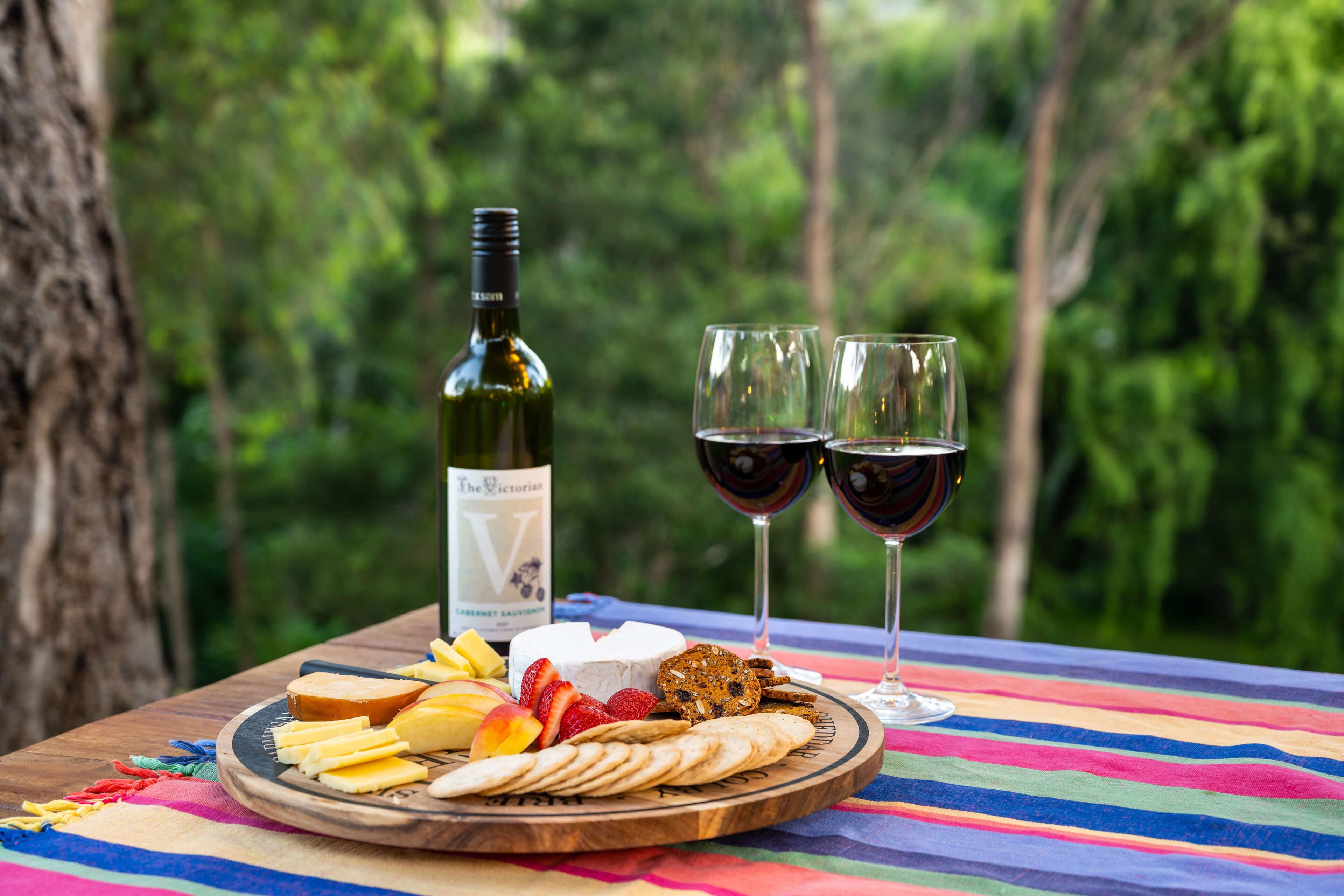 A wooden board with cheese, fruits, crackers, and a bottle of wine on a colorful tablecloth, set against a forest backdrop.