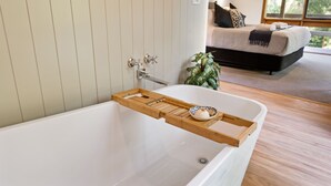 Casa de banho moderna com bandeja de madeira na banheira, uma pequena planta e vista para um quarto com cama e janela.