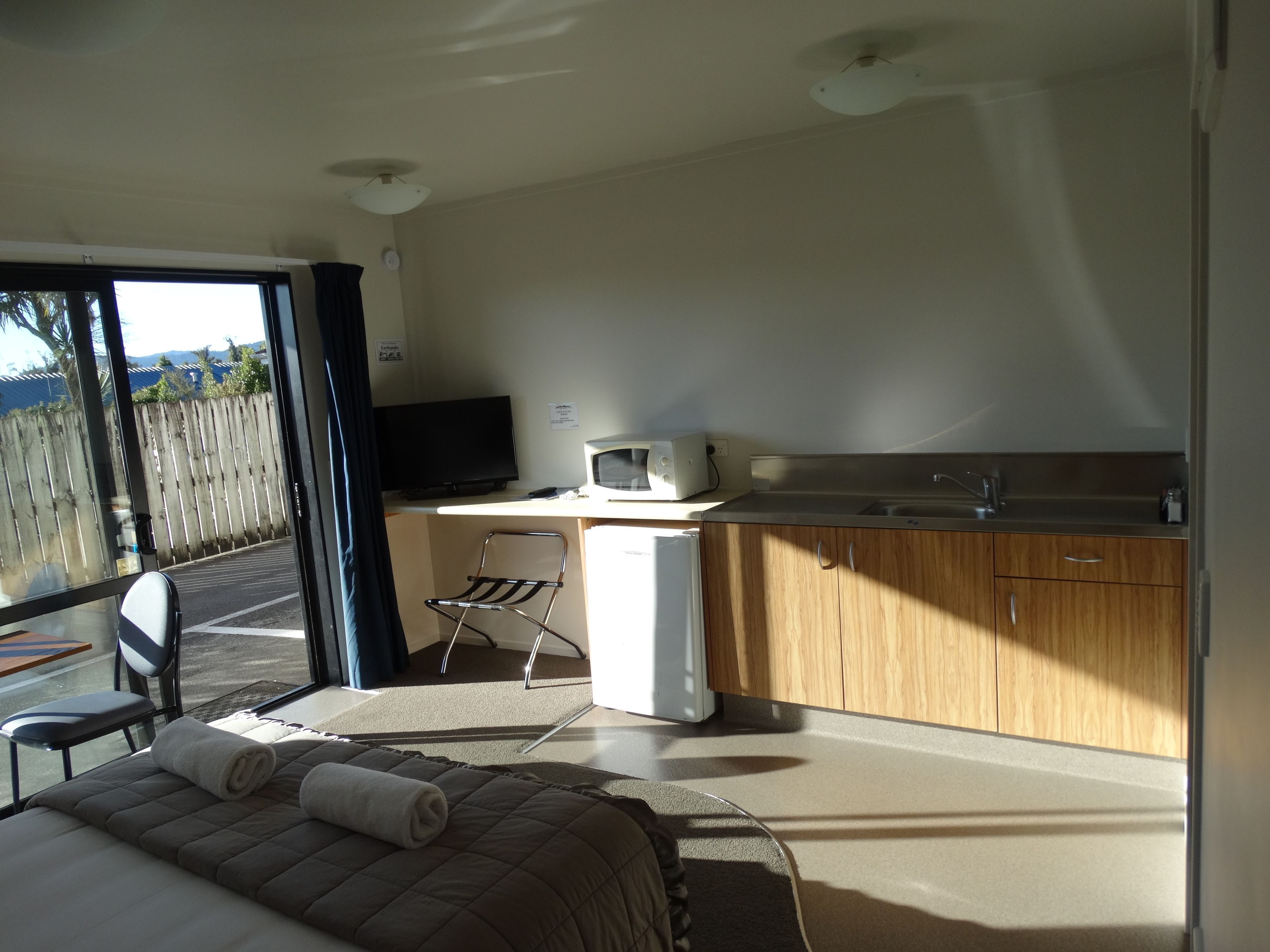 Habitación moderna con cama, escritorio con computadora y microondas, pequeña cocina y vistas a través de un ventanal grande.