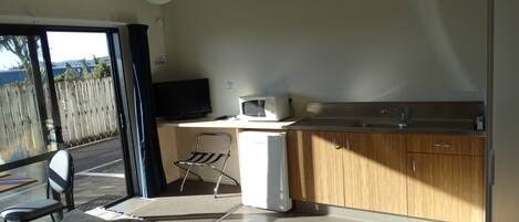 Habitación moderna con cama, escritorio con computadora y microondas, pequeña cocina y vistas a través de un ventanal grande.
