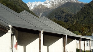 A building with a sloped roof, white walls, and a view of snow-capped mountains in the background. - Alpine Glacier Motel (Franz Josef Glacier)