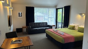 A hotel room with a bed, a sofa, a table with a remote control, and a TV mounted on the wall. - Alpine Glacier Motel (Franz Josef Glacier)