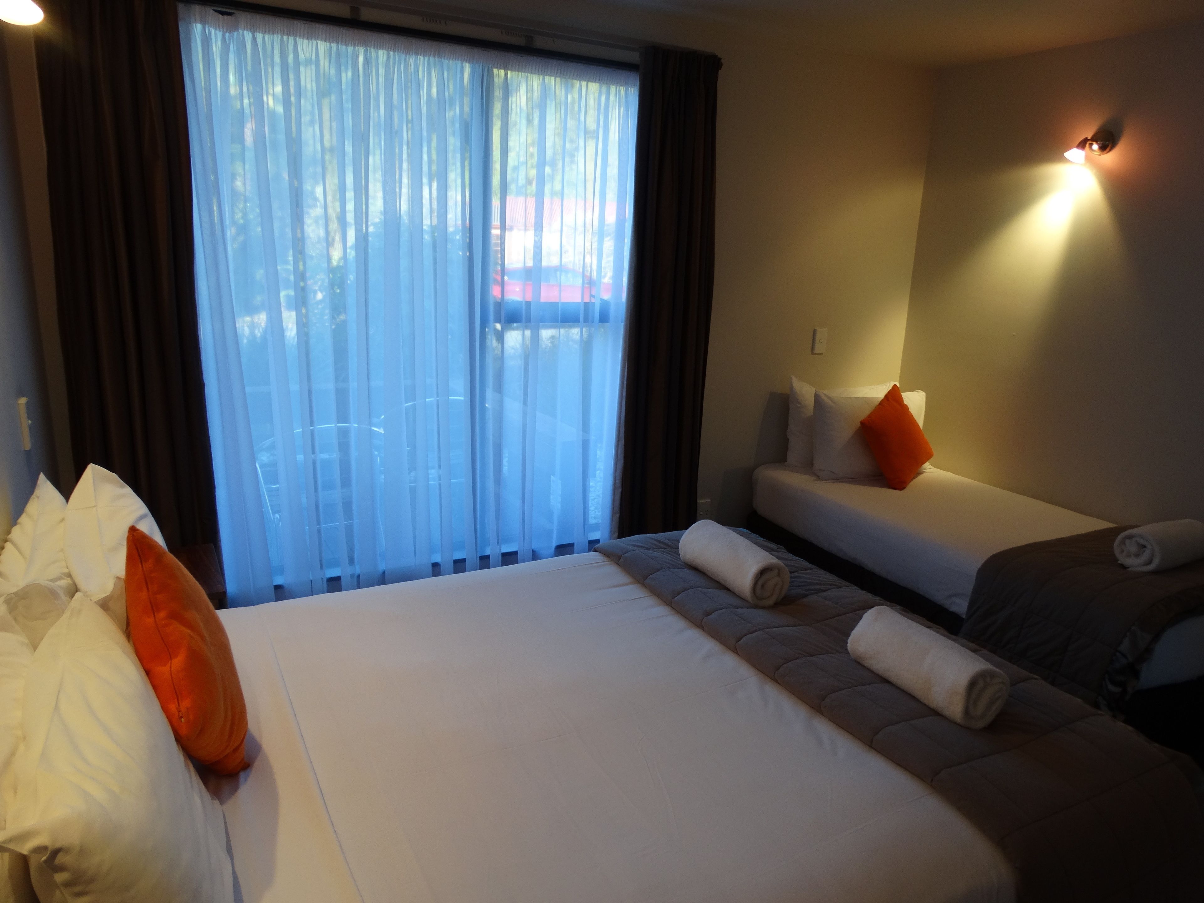 A hotel room with a large bed, a smaller bed, and a sofa. There is a window with curtains, a wall-mounted light, and a view of the outside.