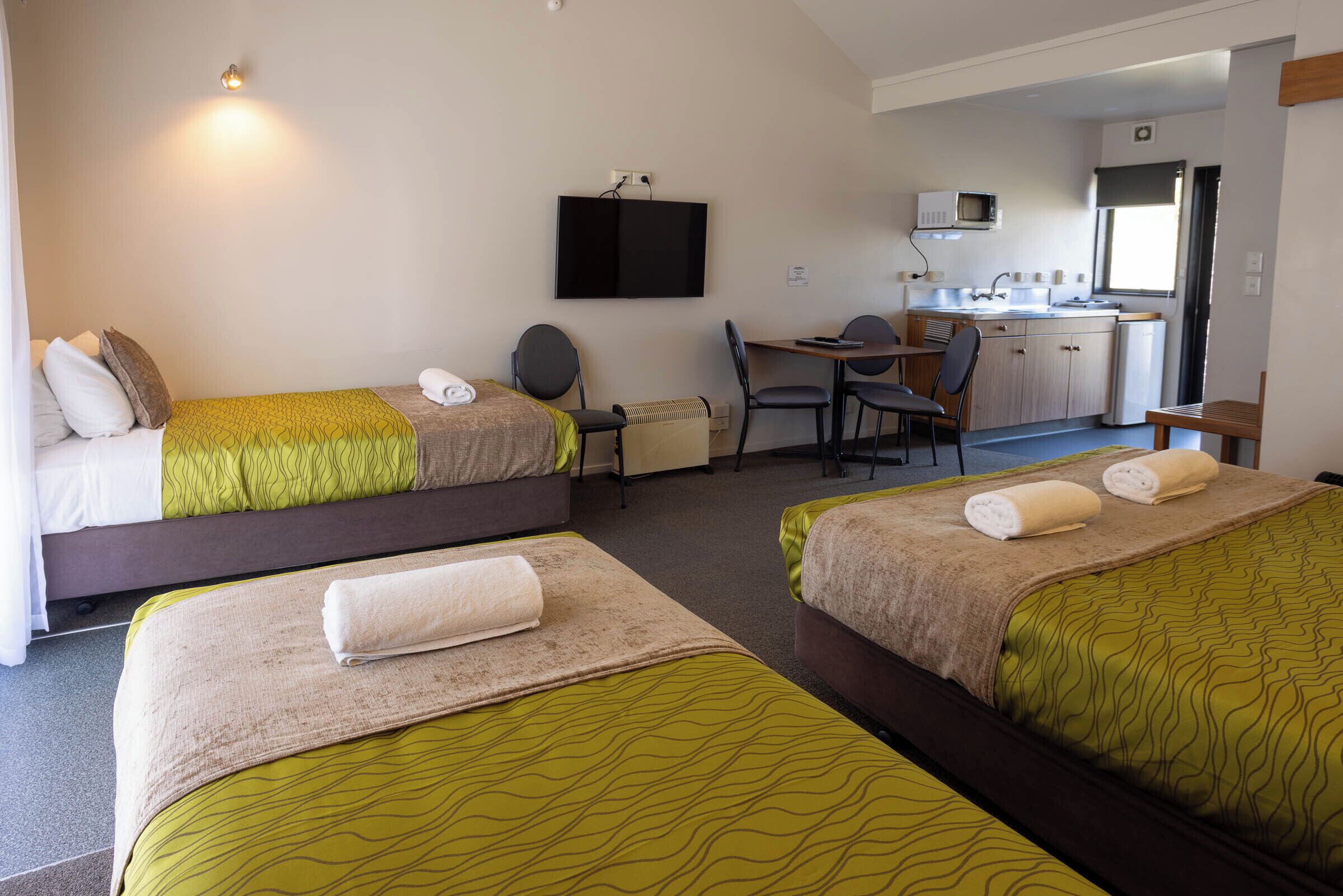 A hotel room with two beds, a desk, a chair, a TV, and a kitchenette.