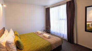 A hotel room with a bed, pillows, a window with curtains, a chair, and a wall-mounted mirror.