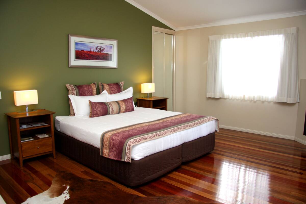A large bed with a brown headboard, two bedside tables, a nightstand, and a framed picture on the wall.