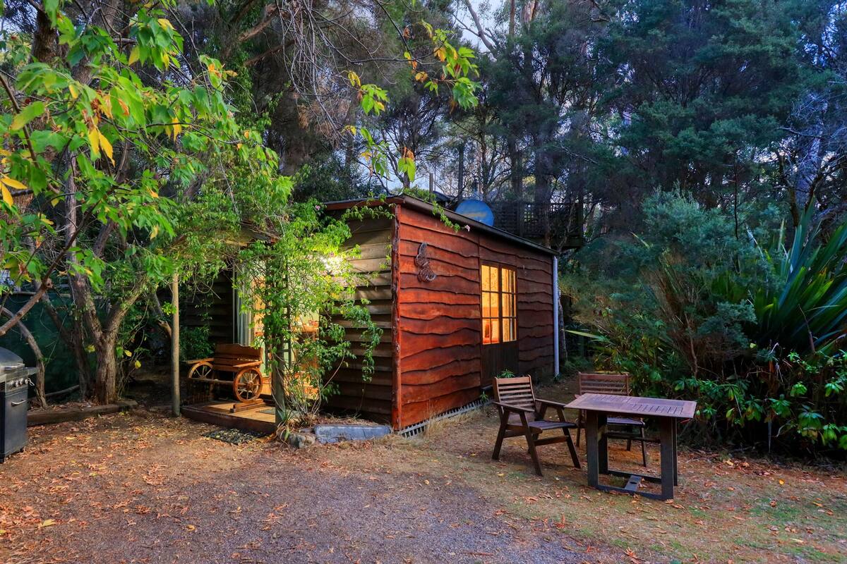 Un chalet rustique en bois, doté d’une véranda et entouré d’une végétation luxuriante et d’arbres.