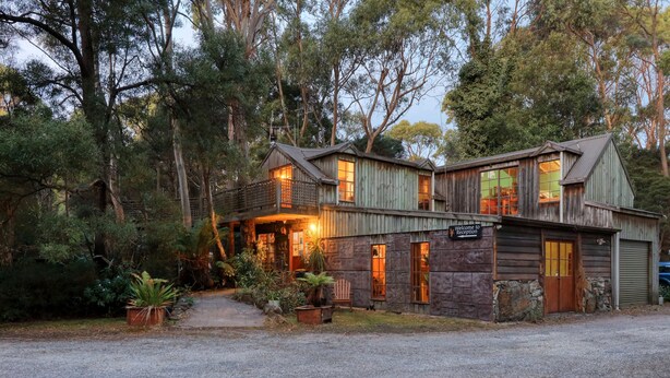 Une maison en bois au style rustique, dotée d’un espace d’accueil et entourée d’arbres denses, avec une allée gravillonnée.